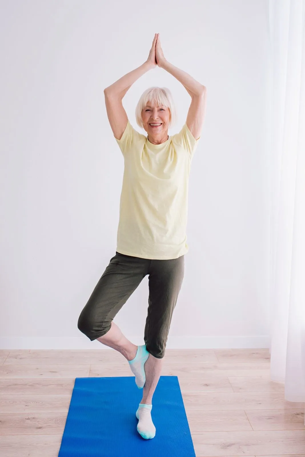 A smiling older woman balancing in tree pose on a mat, hands together overhead