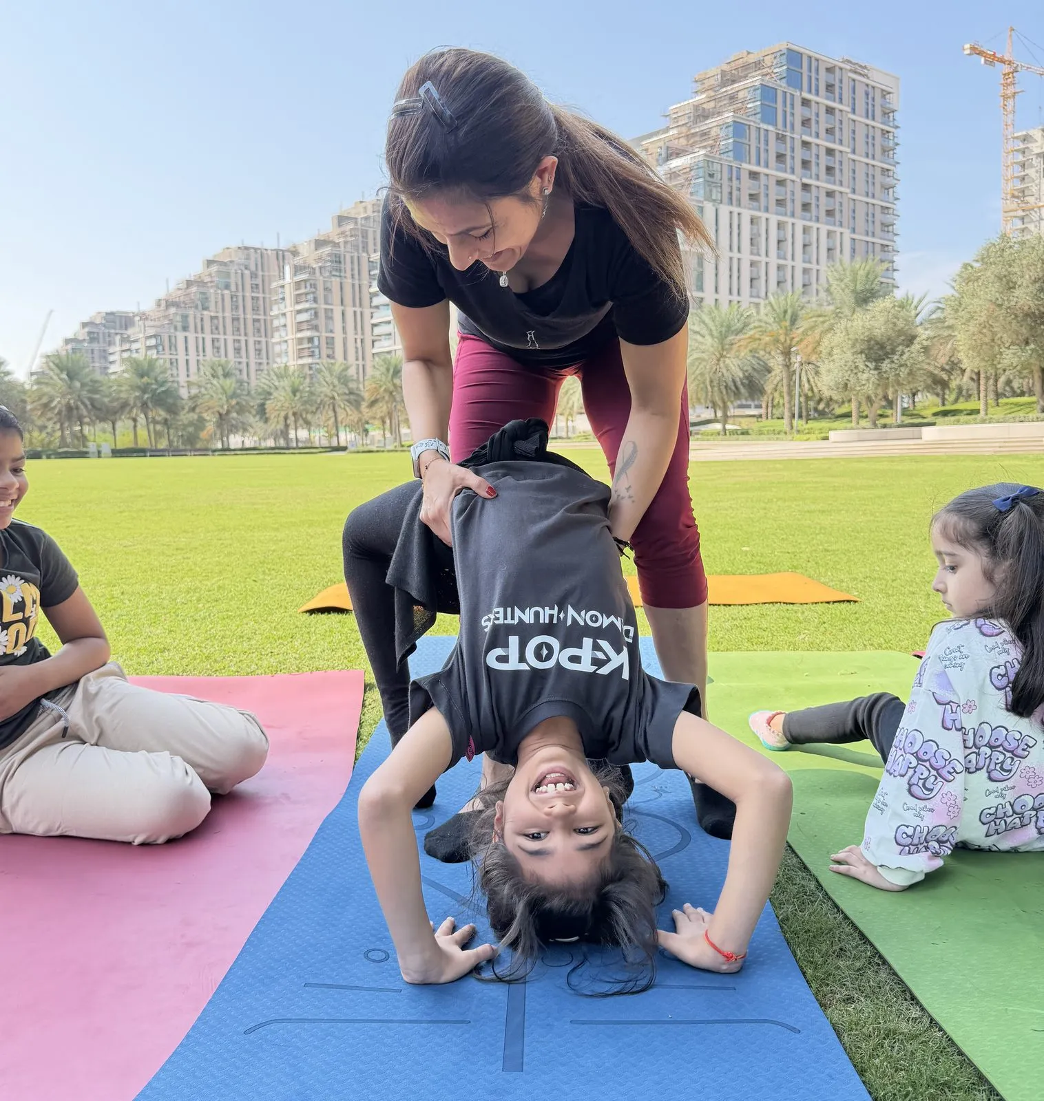 Brinda supporting a child in a shoulder stand while other children watch from their mats in a Dubai park